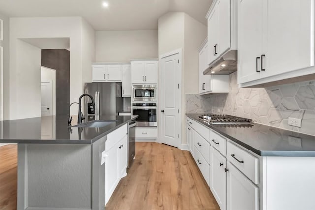 kitchen with stainless steel appliances, under cabinet range hood, dark countertops, a sink, and light wood-type flooring