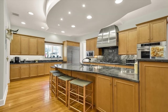 3208 Rain Dance Cv, Austin, TX 78746 - kitchen featuring appliances with stainless steel finishes, custom range hood, decorative backsplash, light wood-style flooring, and a kitchen breakfast bar