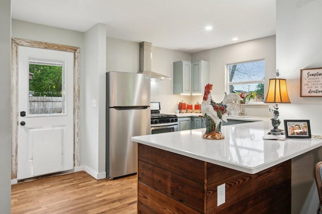 kitchen with light countertops, a peninsula, wall chimney exhaust hood, stainless steel appliances, and a wealth of natural light