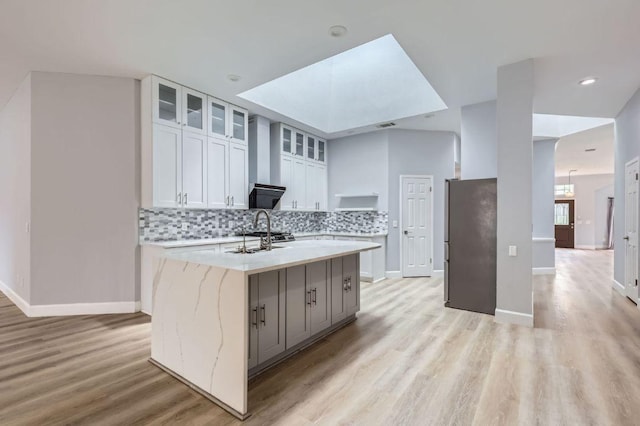kitchen featuring backsplash, glass insert cabinets, a kitchen island with sink, baseboards, and light wood-style floors
