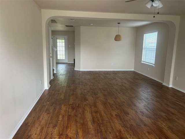 empty room featuring arched walkways, baseboards, a ceiling fan, and dark wood finished floors
