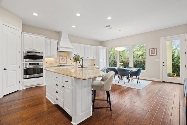 kitchen with custom exhaust hood, a breakfast bar, double oven, white cabinets, and light stone countertops