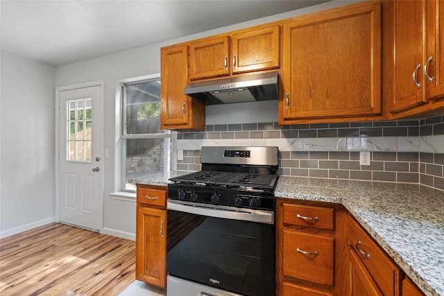 kitchen featuring light wood-style flooring, under cabinet range hood, stainless steel gas range oven, brown cabinetry, and backsplash