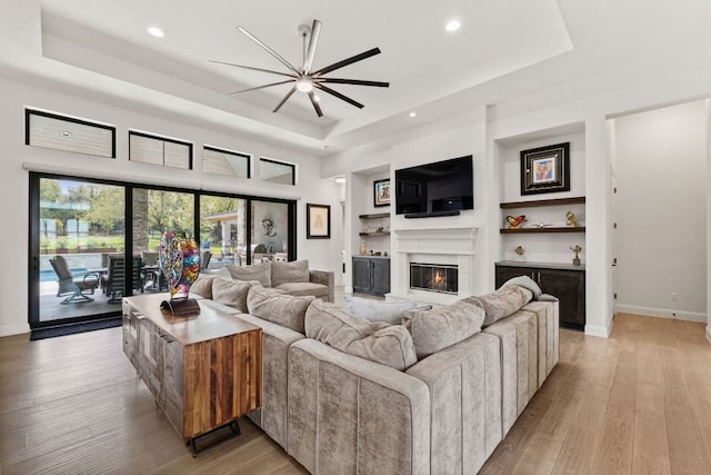 living area with a glass covered fireplace, baseboards, light wood-style floors, a tray ceiling, and ceiling fan