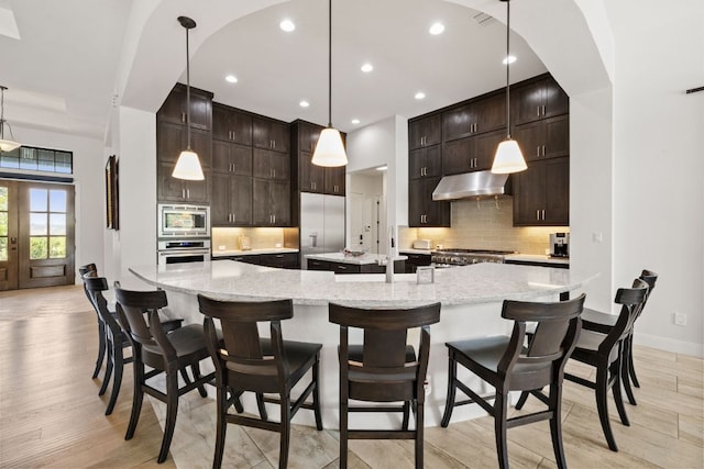 kitchen featuring light wood-style flooring, under cabinet range hood, appliances with stainless steel finishes, decorative backsplash, and a spacious island