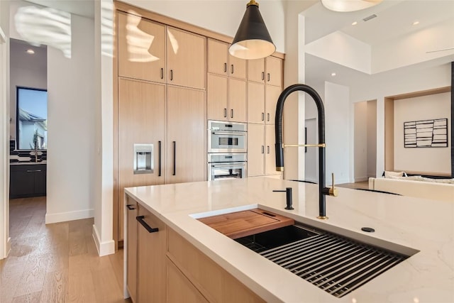 kitchen featuring a sink, light brown cabinetry, light wood finished floors, hanging light fixtures, and light stone counters