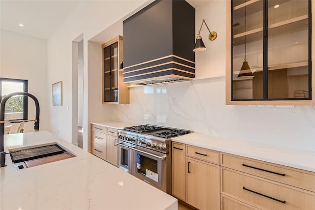 kitchen featuring custom exhaust hood, range with two ovens, light brown cabinetry, decorative backsplash, and a sink