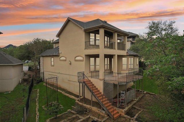 back of property at dusk with fence, a balcony, a yard, stucco siding, and roof with shingles