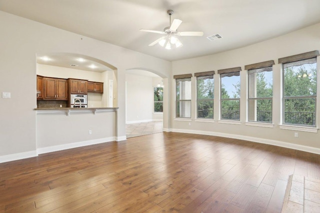 unfurnished living room with hardwood / wood-style floors, ceiling fan, arched walkways, baseboards, and visible vents