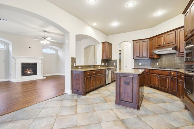 kitchen featuring under cabinet range hood, stainless steel appliances, a ceiling fan, a kitchen island, and decorative backsplash