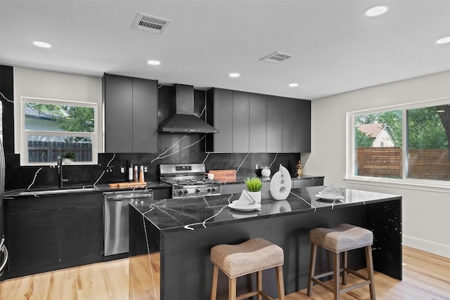 6303 Bexton Cir, Austin, TX 78745 - kitchen featuring wall chimney exhaust hood, appliances with stainless steel finishes, a kitchen island, a breakfast bar, and light wood-type flooring
