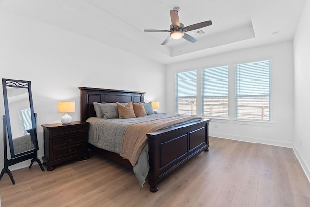 bedroom featuring light wood finished floors, a tray ceiling, baseboards, visible vents, and ceiling fan