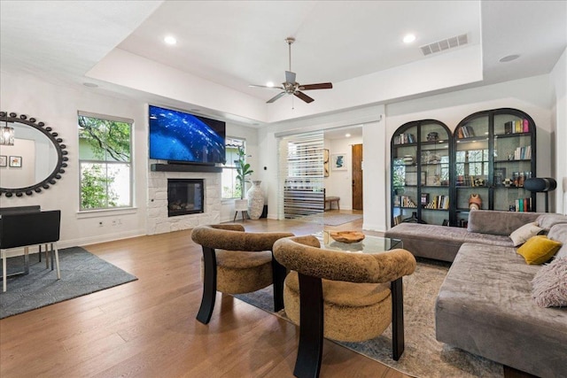 living room with wood finished floors, ceiling fan, a stone fireplace, visible vents, and a tray ceiling