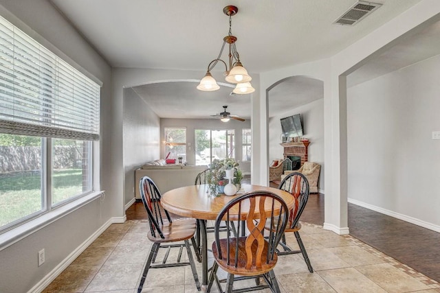 dining space featuring visible vents, light tile patterned floors, arched walkways, a brick fireplace, and ceiling fan with notable chandelier