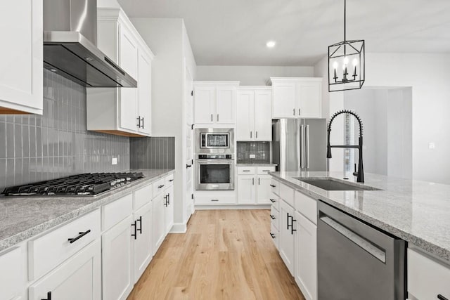 kitchen with light wood finished floors, a sink, a chandelier, wall chimney exhaust hood, and appliances with stainless steel finishes