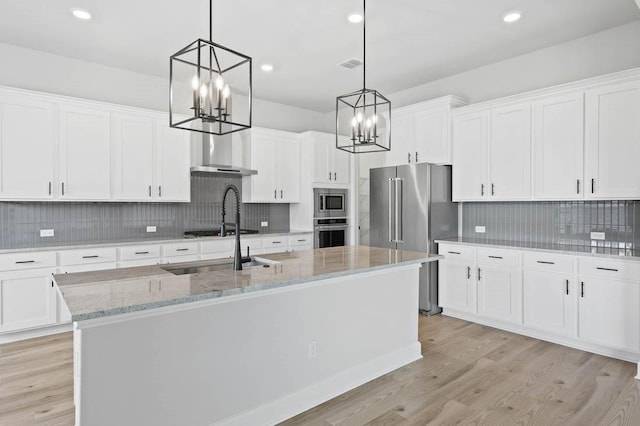 kitchen with appliances with stainless steel finishes, light wood finished floors, a chandelier, decorative backsplash, and an island with sink