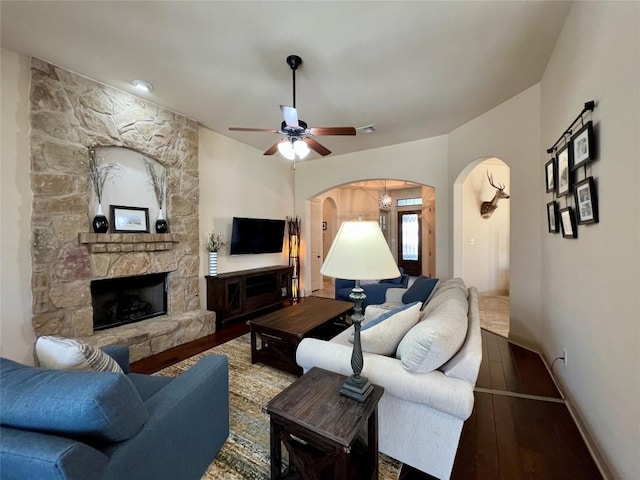 living room with arched walkways, a stone fireplace, hardwood / wood-style floors, a ceiling fan, and baseboards