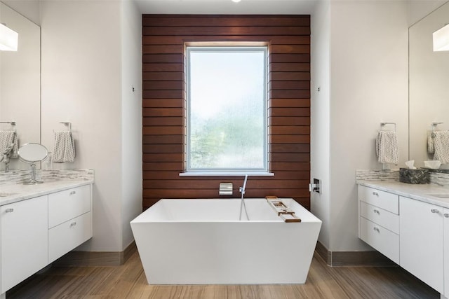 bathroom featuring two vanities, wood finished floors, a freestanding tub, and baseboards