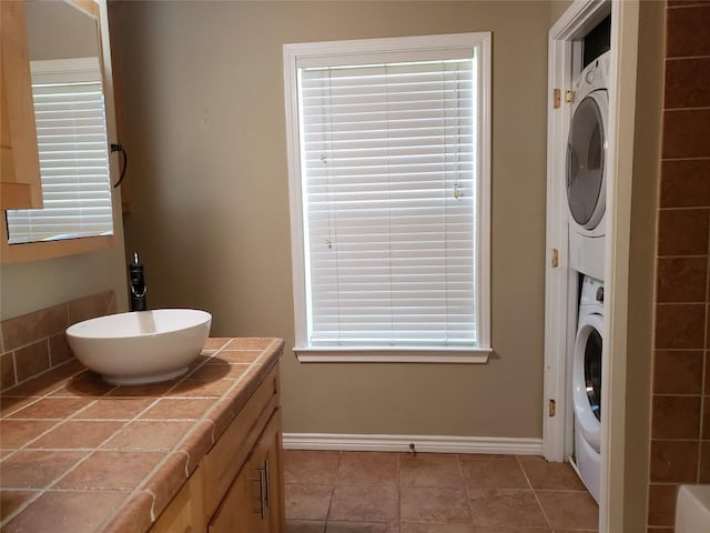 clothes washing area with baseboards, light tile patterned flooring, stacked washing maching and dryer, laundry area, and a sink