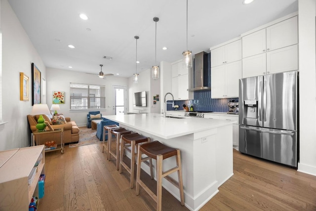 kitchen featuring light wood-style floors, wall chimney range hood, stainless steel refrigerator with ice dispenser, a sink, and decorative backsplash