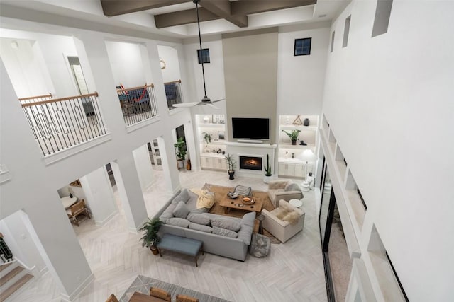 living room featuring ceiling fan, beamed ceiling, a warm lit fireplace, and a towering ceiling