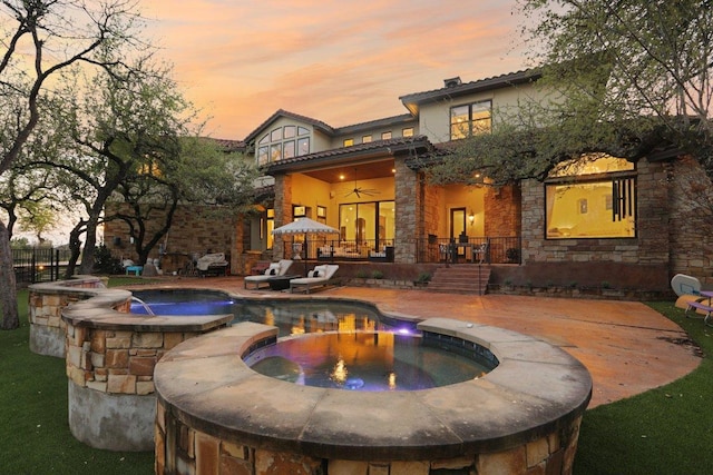 back of house at dusk with a pool with connected hot tub, ceiling fan, a patio, and stone siding