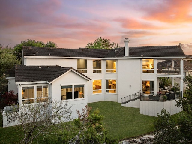 back of property at dusk with a lawn, stucco siding, a balcony, and a sunroom