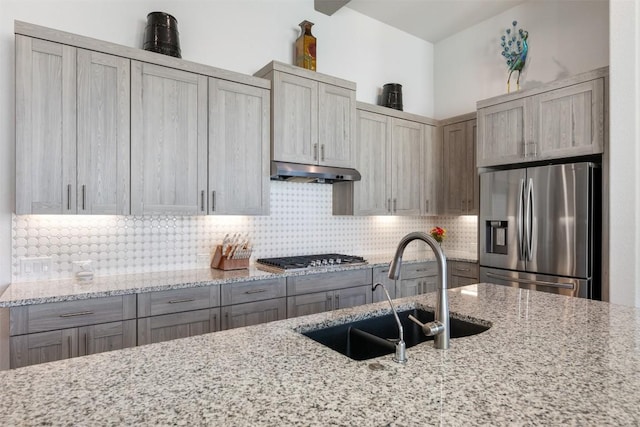 kitchen featuring backsplash, under cabinet range hood, light stone countertops, stainless steel appliances, and a sink
