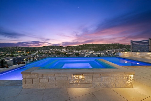 pool at dusk featuring a patio, an infinity pool, an in ground hot tub, and fence