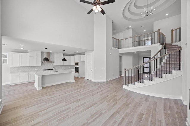unfurnished living room featuring ceiling fan with notable chandelier, light wood-type flooring, a towering ceiling, recessed lighting, and stairs