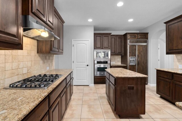 kitchen with built in appliances, dark brown cabinets, under cabinet range hood, arched walkways, and backsplash