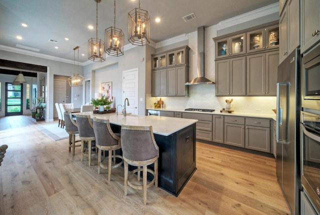 kitchen featuring wall chimney exhaust hood, ornamental molding, appliances with stainless steel finishes, an island with sink, and a kitchen bar