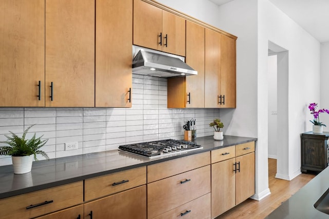 kitchen featuring under cabinet range hood, stainless steel gas stovetop, light wood-style flooring, dark countertops, and backsplash