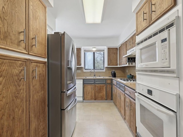 kitchen featuring a sink, brown cabinets, decorative backsplash, under cabinet range hood, and white appliances