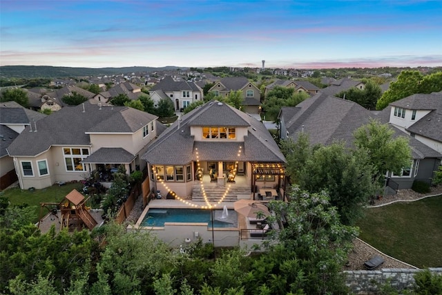 back of house at dusk with a fenced backyard, a patio, an outdoor pool, a shingled roof, and a residential view