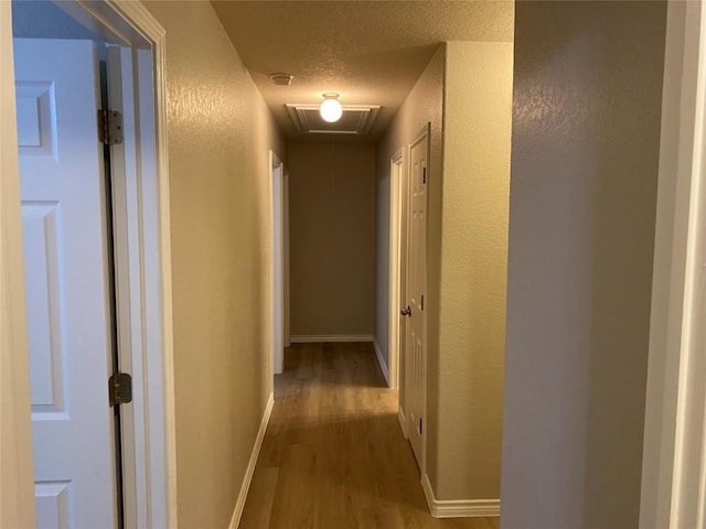 hall featuring a textured wall, baseboards, attic access, and wood finished floors
