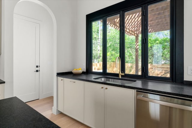 kitchen with stainless steel dishwasher, a sink, dark countertops, light wood-style flooring, and white cabinetry