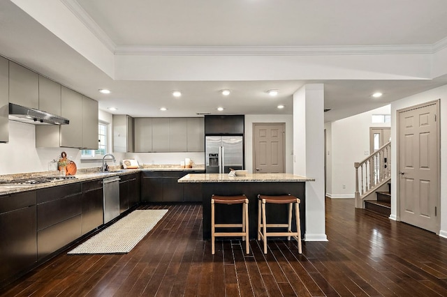 kitchen with under cabinet range hood, stainless steel appliances, a sink, crown molding, and a breakfast bar area