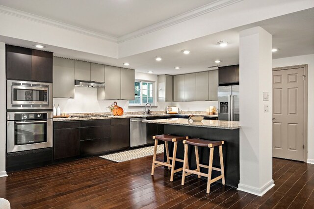kitchen with exhaust hood, dark wood finished floors, a kitchen bar, ornamental molding, and appliances with stainless steel finishes