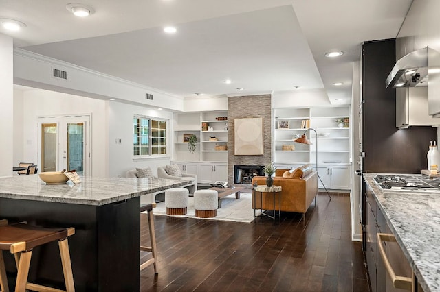 kitchen with wall chimney range hood, dark wood-style floors, stainless steel gas stovetop, a breakfast bar, and visible vents