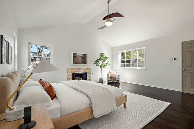bedroom featuring dark wood finished floors, vaulted ceiling, a stone fireplace, baseboards, and ceiling fan
