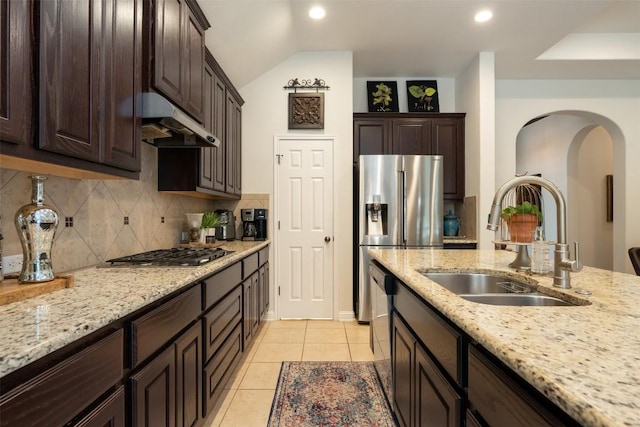 kitchen featuring dark brown cabinetry, backsplash, under cabinet range hood, arched walkways, and a sink