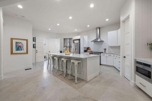 kitchen with recessed lighting, white cabinets, a kitchen island with sink, wall chimney exhaust hood, and appliances with stainless steel finishes