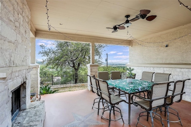 view of patio with outdoor dining area, an outdoor stone fireplace, fence, and ceiling fan