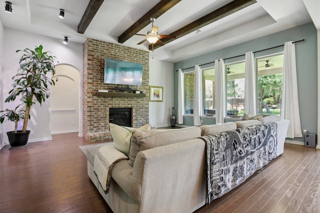 living room with beamed ceiling, a brick fireplace, a ceiling fan, and dark wood-style flooring