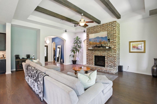 living room featuring a ceiling fan, arched walkways, a brick fireplace, beam ceiling, and dark wood-style floors