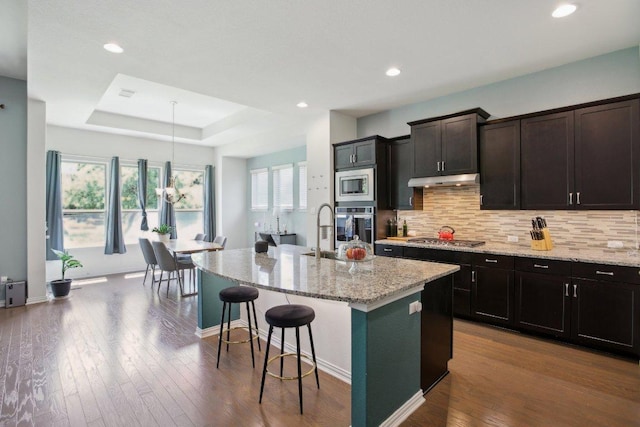 kitchen featuring a kitchen breakfast bar, under cabinet range hood, light stone counters, a kitchen island with sink, and appliances with stainless steel finishes