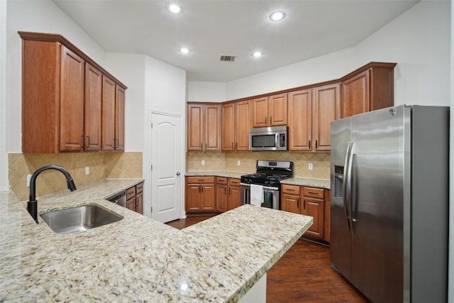 kitchen with brown cabinets, stainless steel appliances, a sink, light stone countertops, and dark wood-style flooring
