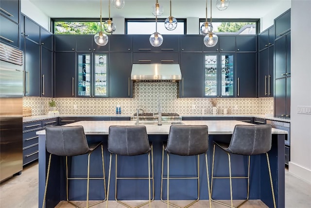 kitchen with under cabinet range hood, stainless steel built in fridge, a center island with sink, tasteful backsplash, and hanging light fixtures