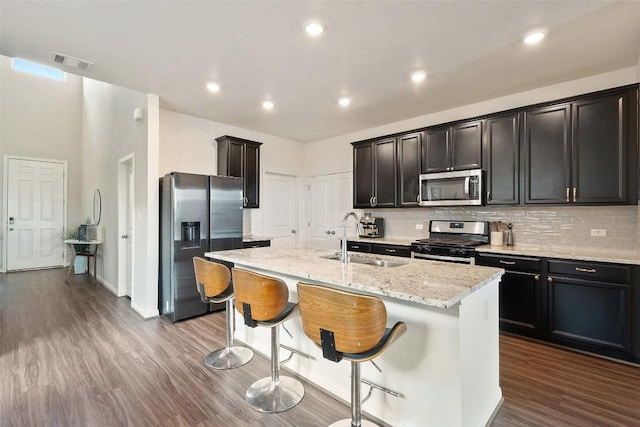 kitchen with appliances with stainless steel finishes, a sink, a kitchen island with sink, light stone counters, and decorative backsplash